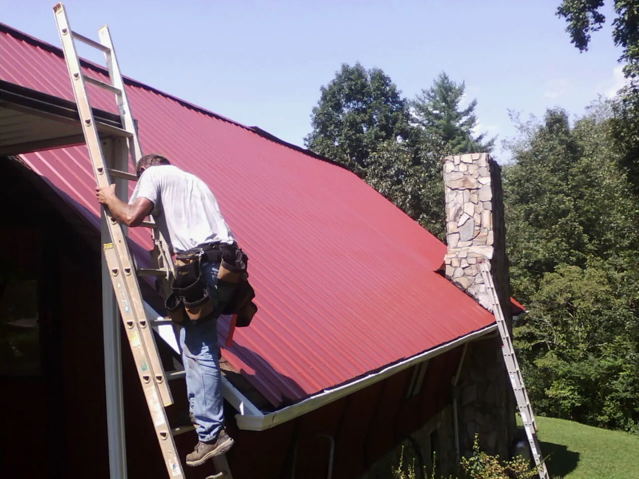 Roofer on a ladder beside a red metal roof and stone chimney — Soffit & Fascia Repair by Roofing Brothers in Fisherville